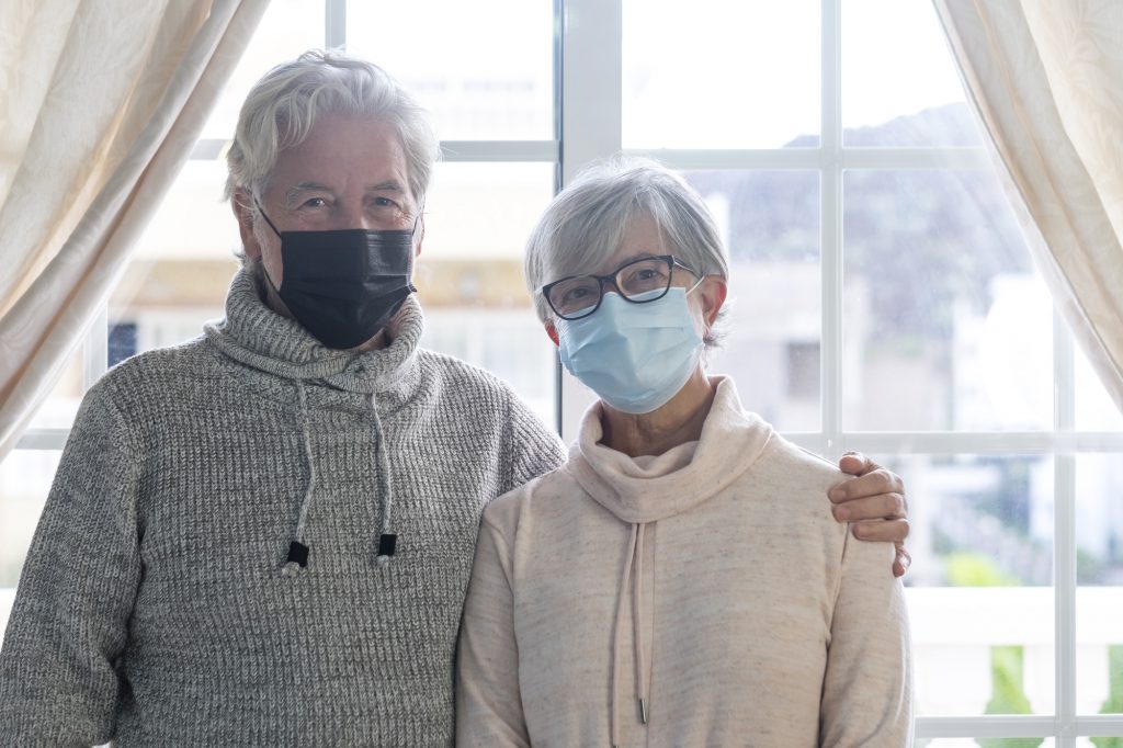 Senior couple look at camera wearing surgical protective mask, in lockdown at home for coronavirus